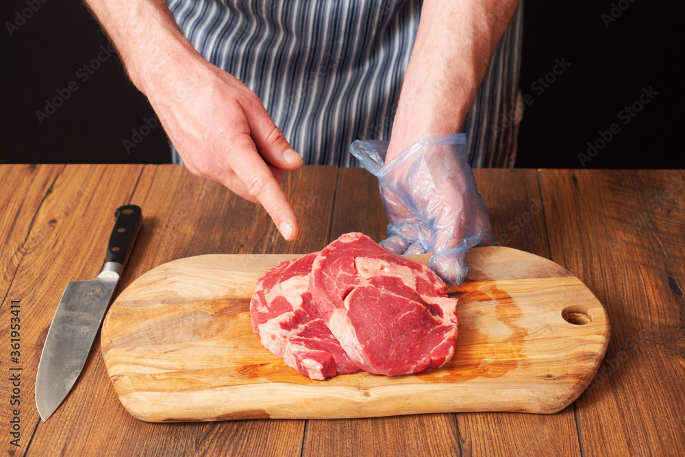 Butcher holding wooden cutting board with two fresh raw rib eye steaks. Right hand pointing to the meat. Knife on the table. Black background.