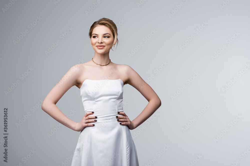cheerful bride in wedding dress standing with hands on hips isolated on grey
