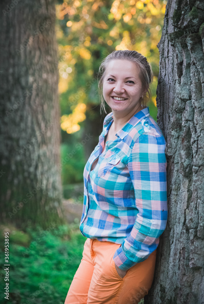 blonde in orange trousers and a plaid shirt standing near a tree in the forest and smiling blonde in orange trousers and a plaid shirt standing near a tree in the forest and smiling