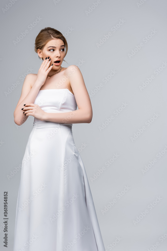 shocked bride in wedding dress looking away and touching face isolated on grey