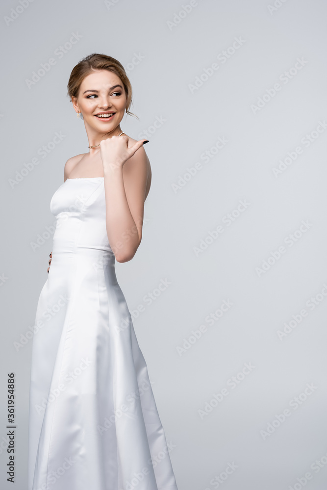 beautiful woman in white wedding dress pointing with thumb isolated on grey