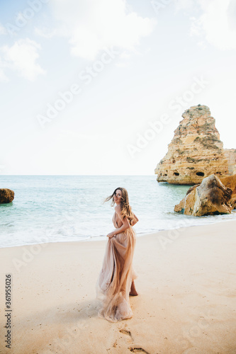stylish girl in a yellow dress is standing with her back with flying hair from the wind at sunset on the beach and the sea