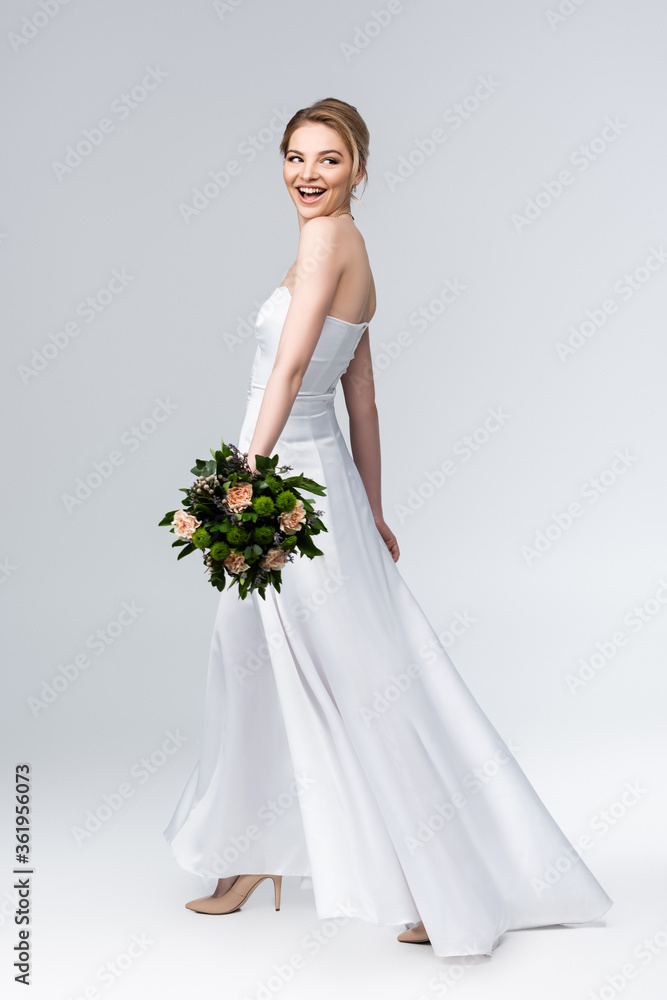 attractive bride in elegant wedding dress holding bouquet of flowers and smiling on grey attractive bride in elegant wedding dress holding bouquet of flowers and smiling on grey