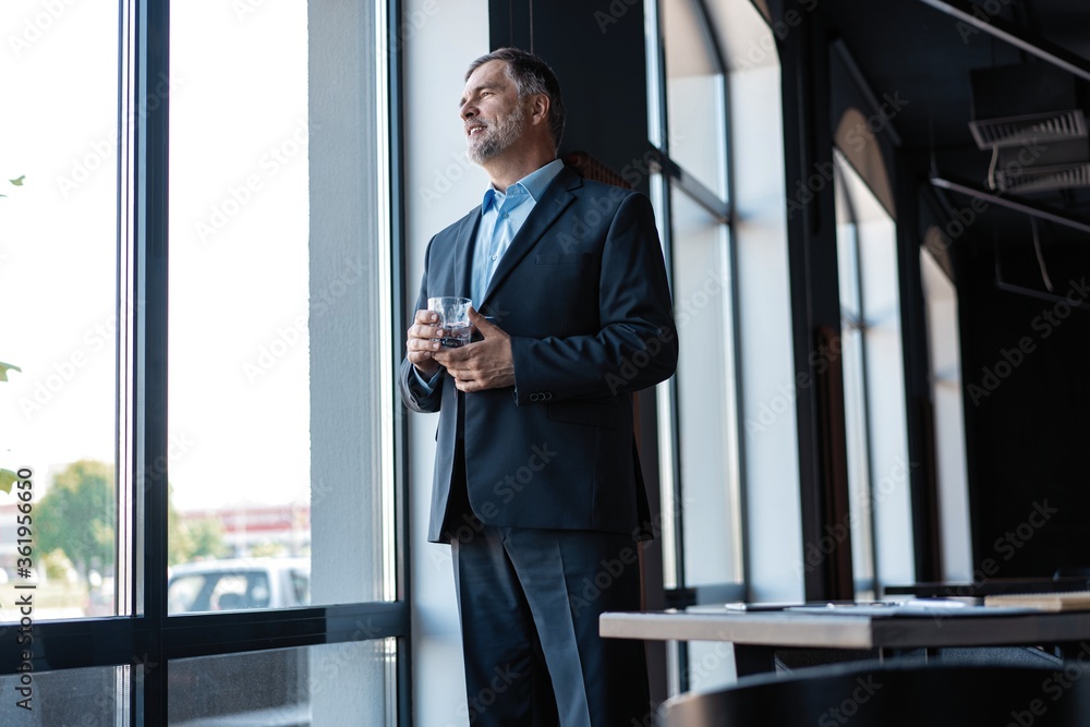 Fototapeta premium Mature businessman drinking water and looking out of a window at the city from an office building.