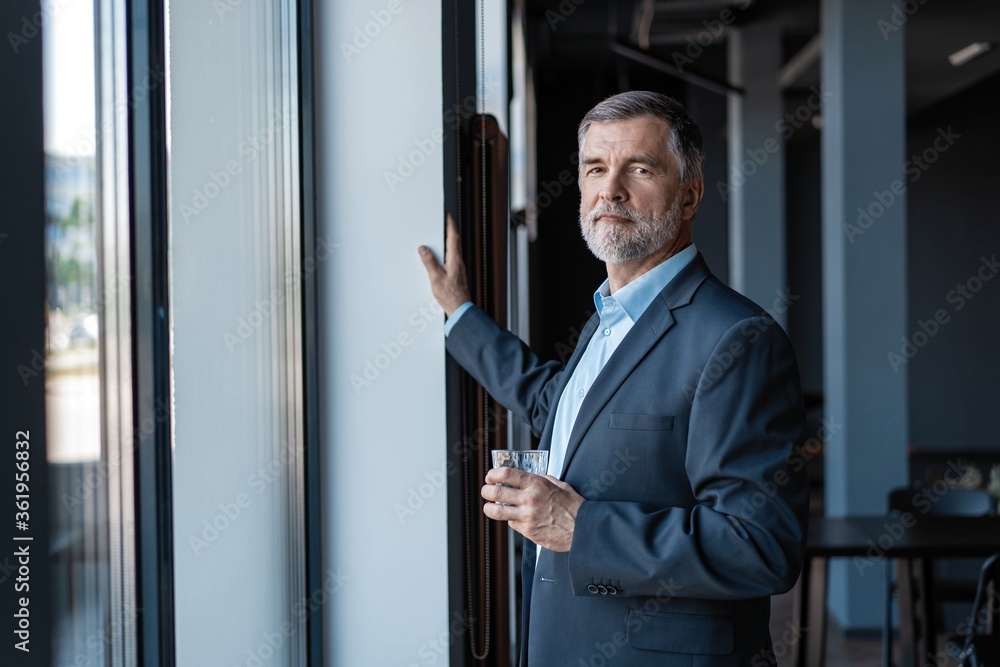 Mature businessman drinking water and looking out of a window at the city from an office building. Mature businessman drinking water and looking out of a window at the city from an office building.