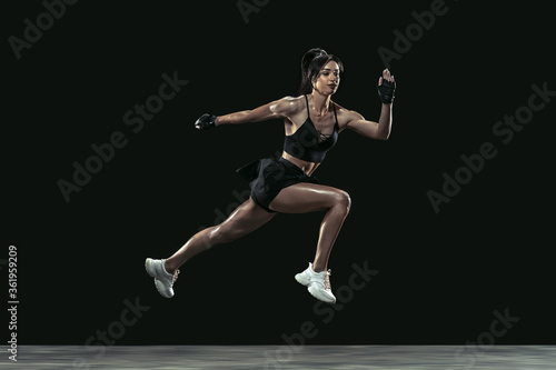 Beautiful young female athlete practicing on black studio background, full length portrait. Sportive fit brunette model in run, jump. Body building, healthy lifestyle, beauty and action concept.