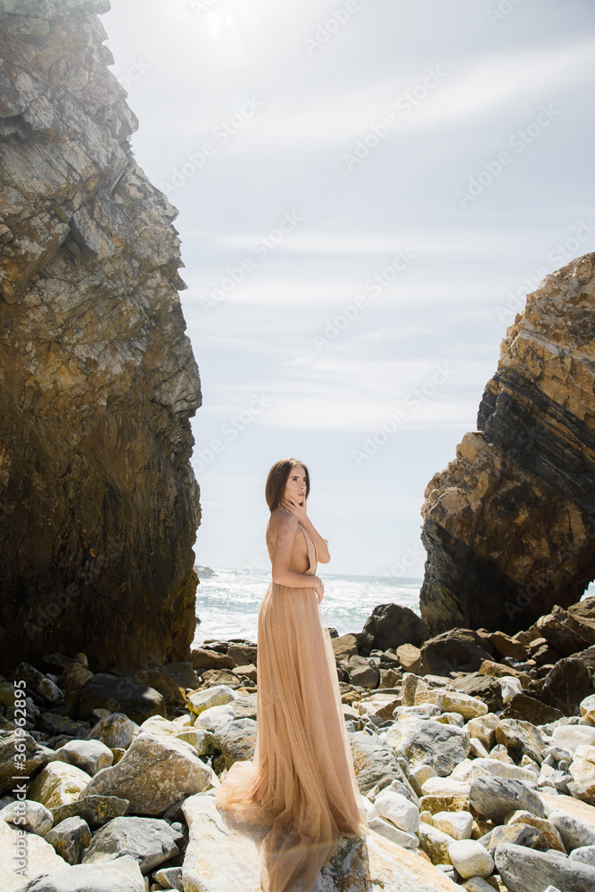 Beautiful girl in a white dress, with long hair and a crown on her head near the sea goes Against the background of rocks