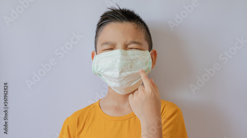 An Asian boy who is closing their eyes wears a COVID-19 virus mask and is dustproof. Raise his hand and point his mask. White background in the house