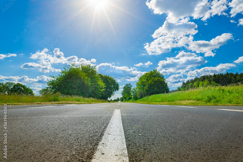 Asphalt road with white dividing lines, landscape and cloudy sky with sun