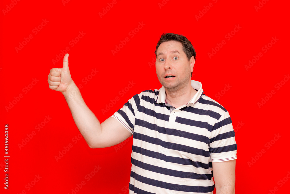 Handsome caucasian man wearing casual striped t-shirt standing over red wall bewildered shows thumb up. Handsome caucasian man wearing casual striped t-shirt standing over red wall bewildered shows thumb up.