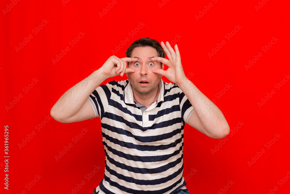 American 30s man wearing casual striped t-shirt standing over isolated red background. Pointing to the eye American 30s man wearing casual striped t-shirt standing over isolated red background. Pointing to the eye
