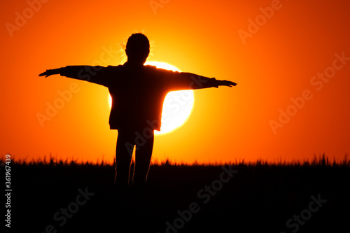 A little girl stands on the meadow and watches the beautiful sunset with her hands outstretched. close up of the sun in the big orange sky.