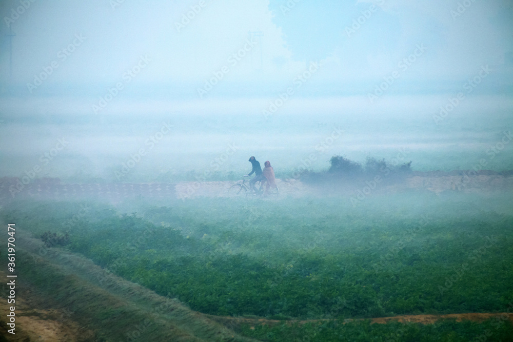 An unidentified man and a woman on a bicycle in early morning with fog around the outskirt area at Agra