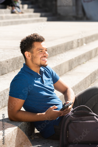 Close up smiling young african american man sitting outside on steps holding cellphone