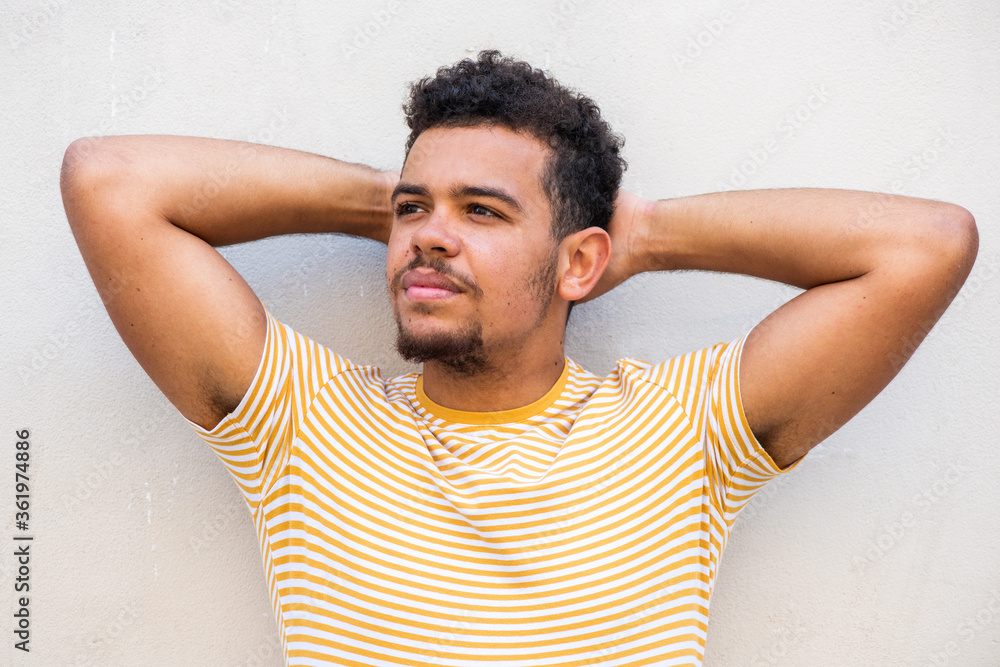 Close up cool young man staring with hands behind head against white wall Close up cool young man staring with hands behind head against white wall
