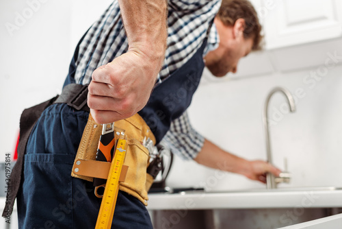 Selective focus of plumber taking wrench from tool belt while fixing faucet in kitchen