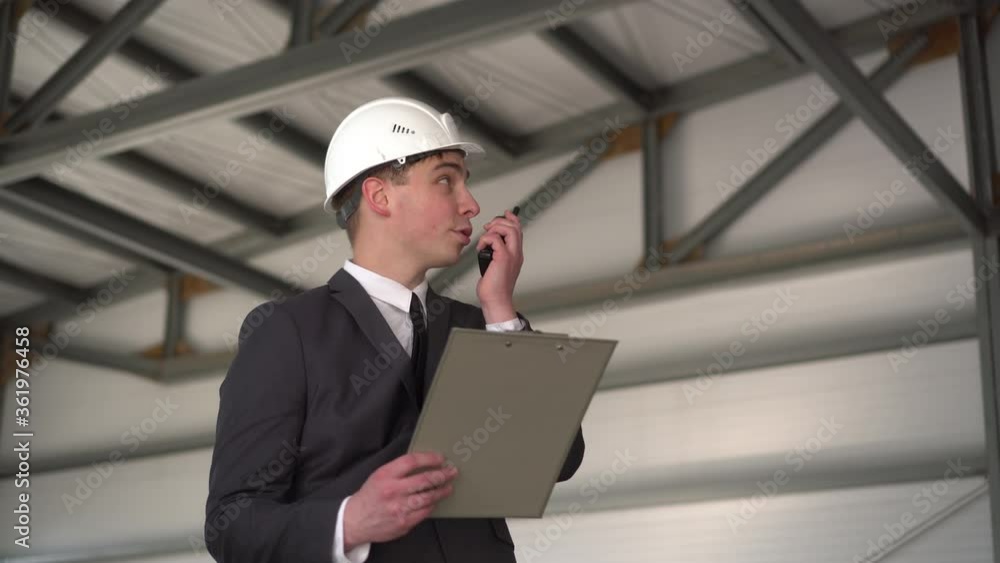 A young man in a helmet with documents speaks on a walkie-talkie at a construction site. The boss in a suit keeps records of documents on architecture. A man looks around. A young man in a helmet with documents speaks on a walkie-talkie at a construction site. The boss in a suit keeps records of documents on architecture. A man looks around.