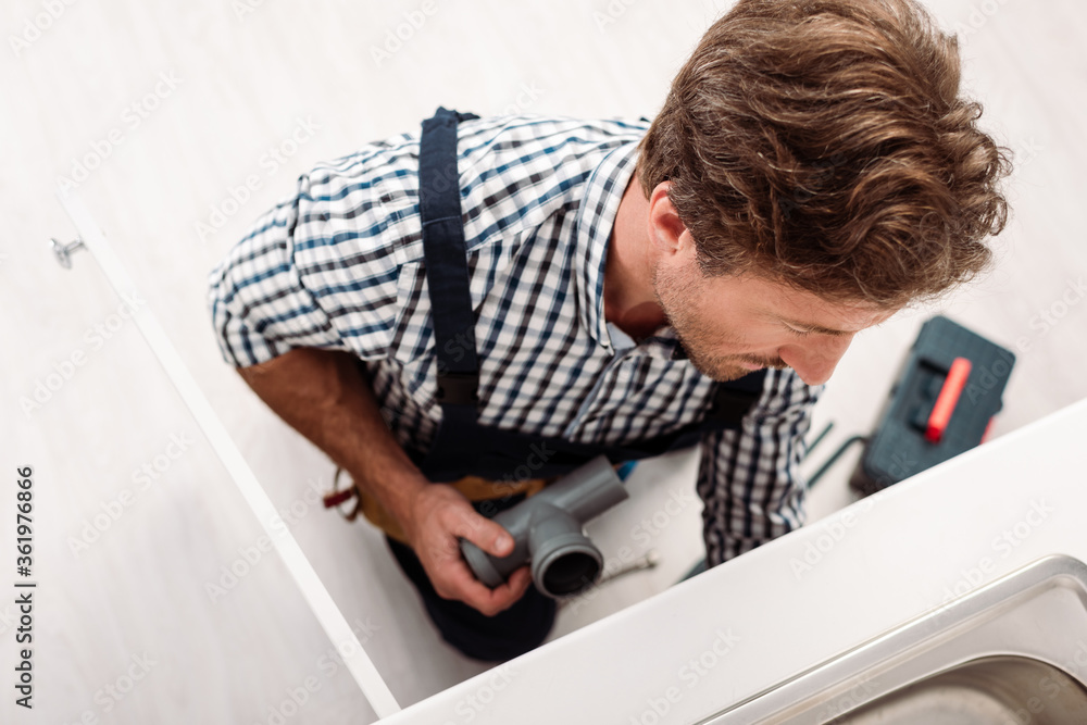 Obraz premium Overhead view of plumber holding plastic pipe while repairing sink near toolbox in kitchen
