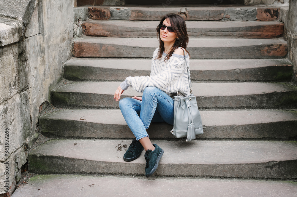 Full-length portrait in autumn weather outdoors, caucasian brunette girl with a slim figure in jeans, a light sweater and with a leather gray backpack. Fashionable model sits on the stairs. Full-length portrait in autumn weather outdoors, caucasian brunette girl with a slim figure in jeans, a light sweater and with a leather gray backpack. Fashionable model sits on the stairs.