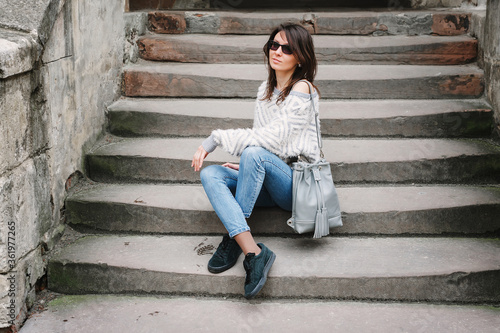 Full-length portrait in autumn weather outdoors, caucasian brunette girl with a slim figure in jeans, a light sweater and with a leather gray backpack. Fashionable model sits on the stairs.