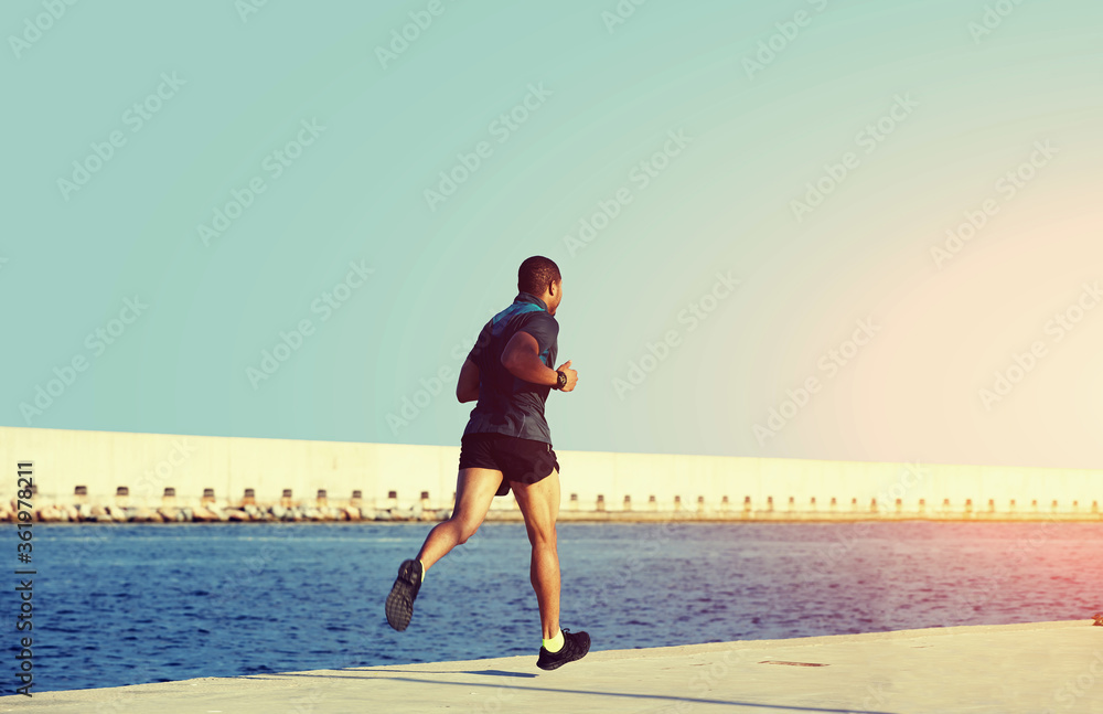 Afro american male runner with muscular body jogging along marina port with sky copy space area for your text message or advertising, sporty young man working out at sunny afternoon outdoors, flare