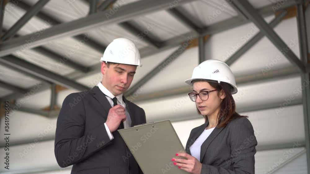 Young man and woman in helmets with documents at a construction site. Chiefs in suits discussing the project of architecture. People look around. Young man and woman in helmets with documents at a construction site. Chiefs in suits discussing the project of architecture. People look around.