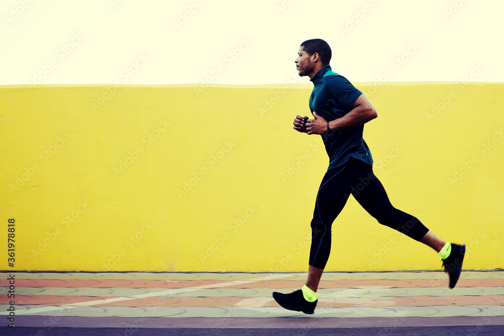 Full length portrait of afro american runner in tracksuit jogging fast against yellow background with copy space area for text message or advertising content, athletic build man working out outdoors Full length portrait of afro american runner in tracksuit jogging fast against yellow background with copy space area for text message or advertising content, athletic build man working out outdoors