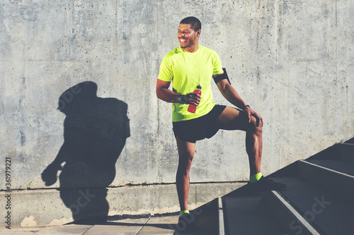Smiling male runner in bright t-shirt holding bottle of energy drink while standing against cement wall background with copy space area for your advertising, happy afro american athlete having a rest