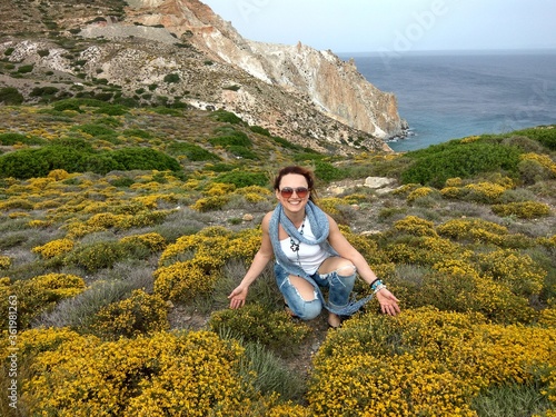 Beautiful girl in glasses and denim pants. Plantation of yellow flowers in the mountains. Against the background of the blue sea. Greece.