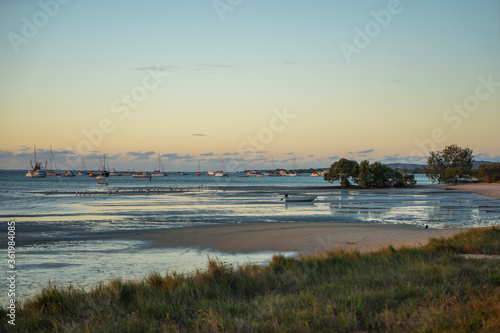 Fotografie Twilight view of beach at low tide with grassy foreground, sand, trees, boats, and clouds on the horizon