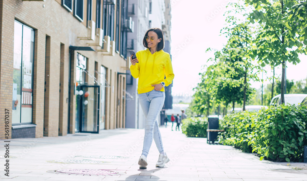 Sunny walk. A full-length photo of a nice smiley girl in a yellow hoodie and glasses on the street, with a white smartwatch on her left hand, looking at her phone.