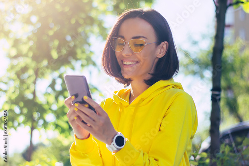 Reading news. A close-up photo of a modern urban girl in a yellow hoodie and glasses, with yellow nails and smartwatch on her hand, watching on her phone.