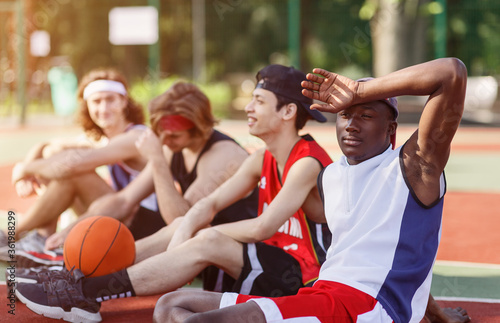 Exhausted black basketball player resting with his diverse team at sports court outside