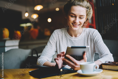 Smiling female in good mood browsing internet websites to do shopping online using modern touch pad connecting to high speed 4G internet enjoying free time with coffee in cafeteria indoors