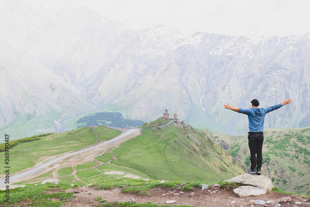 Male tourist in casual clothes with spreaded hands stands on rock looking to Gergeti trinity church surounded by green nature. Travel in Kazbegi national park.