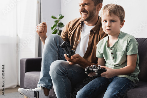 KYIV, UKRAINE - JUNE 9, 2020: upset boy holding joystick and looking at camera near excited father showing winner gesture