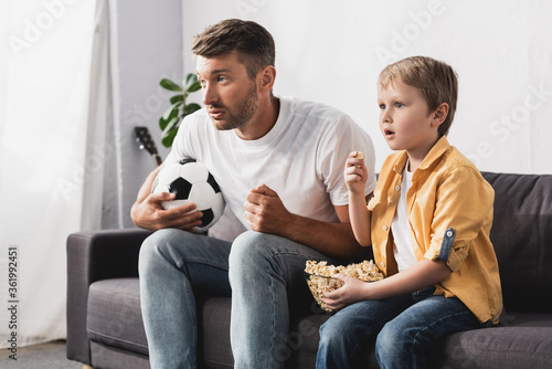 worried father and son watching tv while holding soccer ball and bowl of popcorn