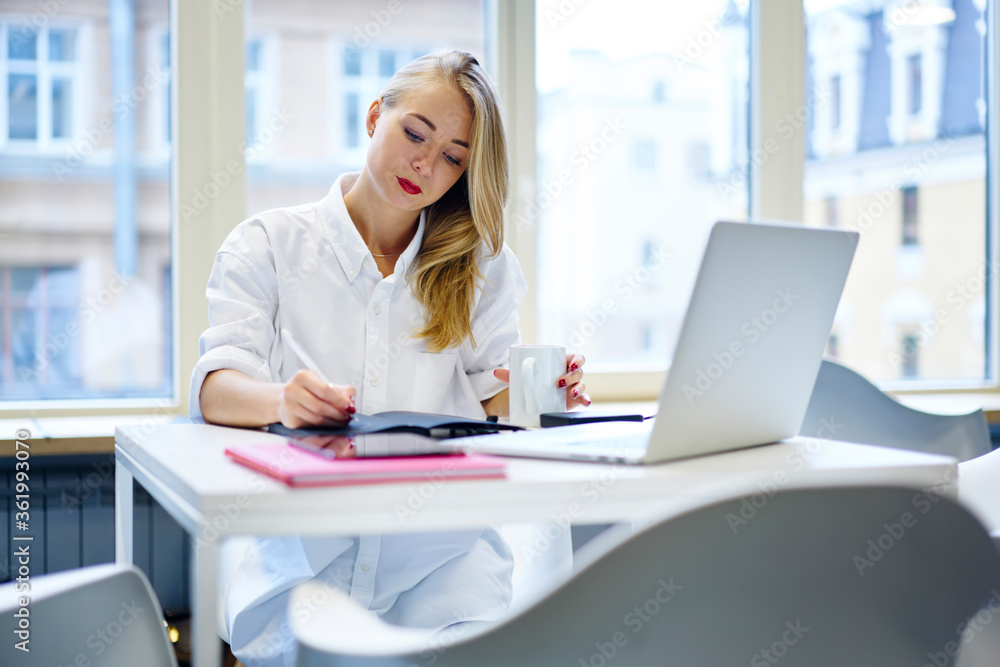 Pensive female entrepreneur making notes of working plans while having coffee break in office, charming administrative manager writing strategy planning using laptop computer for making money transfer Pensive female entrepreneur making notes of working plans while having coffee break in office, charming administrative manager writing strategy planning using laptop computer for making money transfer