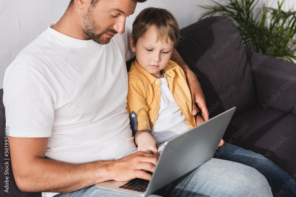 attentive father and son using laptop while sitting on sofa at home