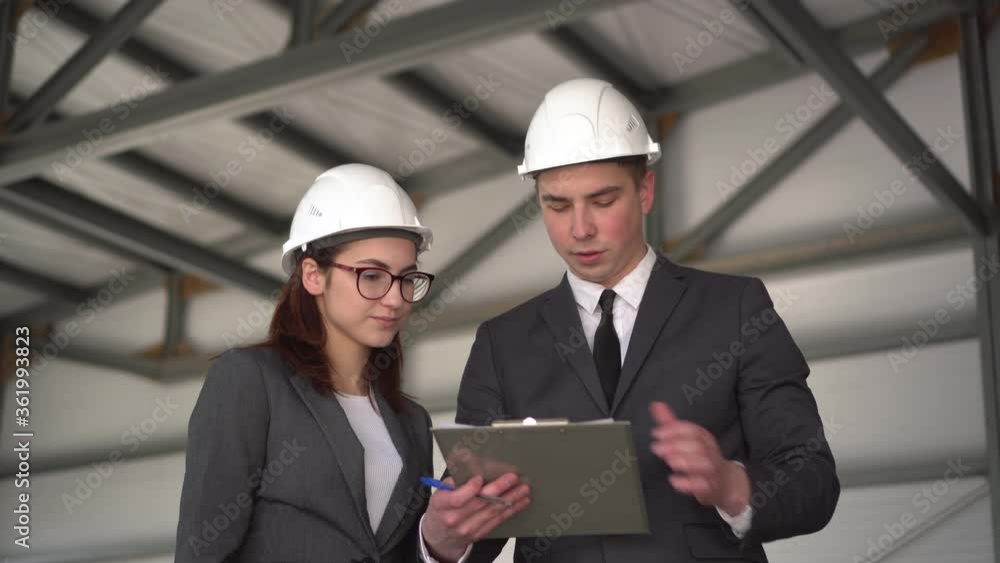 Young man and woman in helmets with documents at a construction site. Bosses in suits are discussing an architecture project. The man is nervous. People look around. Young man and woman in helmets with documents at a construction site. Bosses in suits are discussing an architecture project. The man is nervous. People look around.