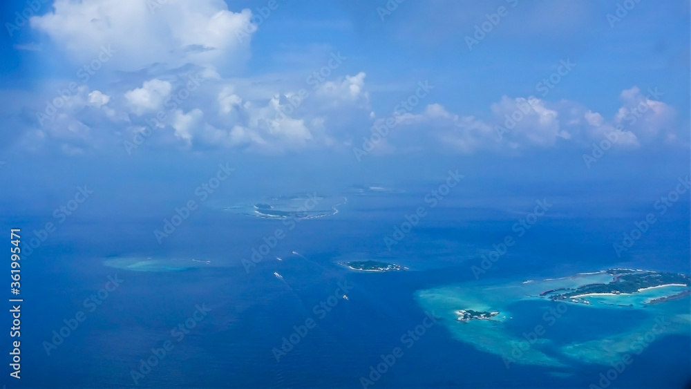 Maldives aerial view. In the middle of the turquoise Indian Ocean are scattered small islands, atolls, with tropical vegetation. Ships are visible on the surface of the water. Azure sky with  clouds.
