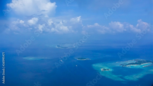 Maldives aerial view. In the middle of the turquoise Indian Ocean are scattered small islands, atolls, with tropical vegetation. Ships are visible on the surface of the water. Azure sky with clouds.