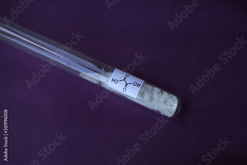 White crystalline substance, organic oxalic acid in a test tube, with a structural formula, on a dark purple background.