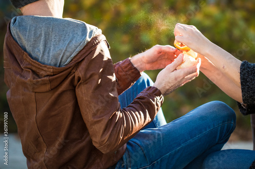 Young couple plays outdoors peeling an orange fruit. Male hands in brown leather jacket hold citrus and woman ready to catch it. Young family outdoors. Sunny day, dark green background out of focus