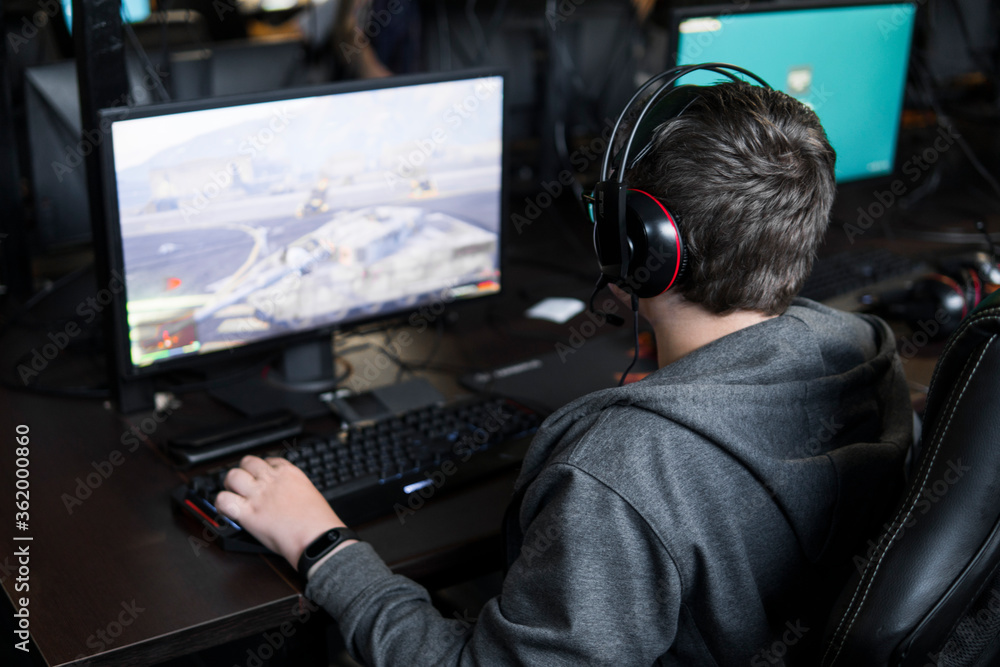 POLAND, WARSAW - June 10, 2020: Back view of young gamer in headphone playing video game at computer club. POLAND, WARSAW - June 10, 2020: Back view of young gamer in headphone playing video game at computer club.