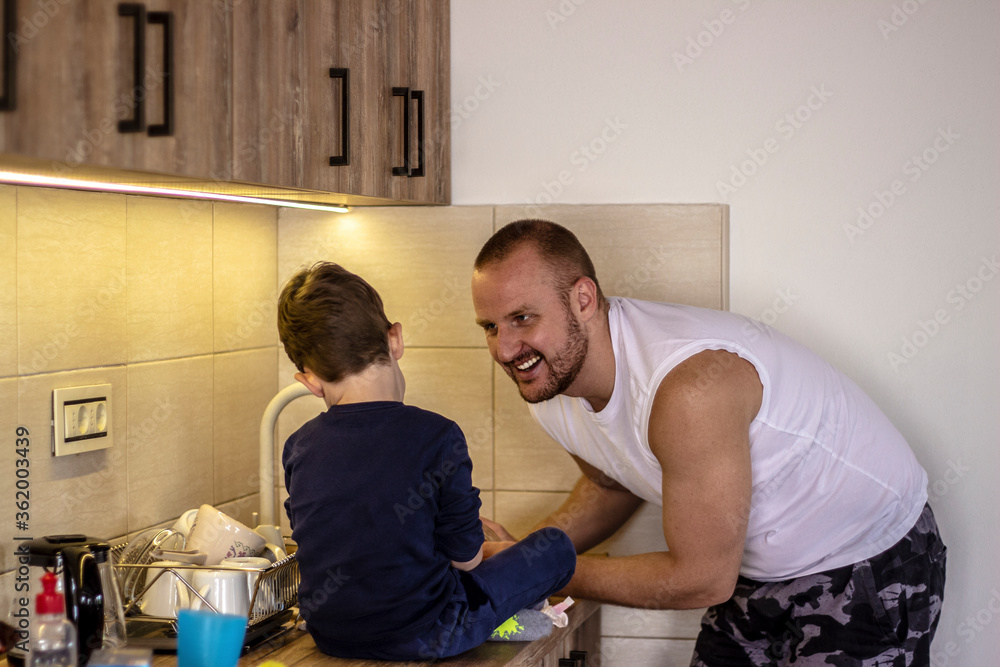 Obraz premium Smilling father looking at his son while cleaning dishes. Son is watching at his father in the kitchen washing dishes. Happy dad and son spend time at home enjoying weekend together.