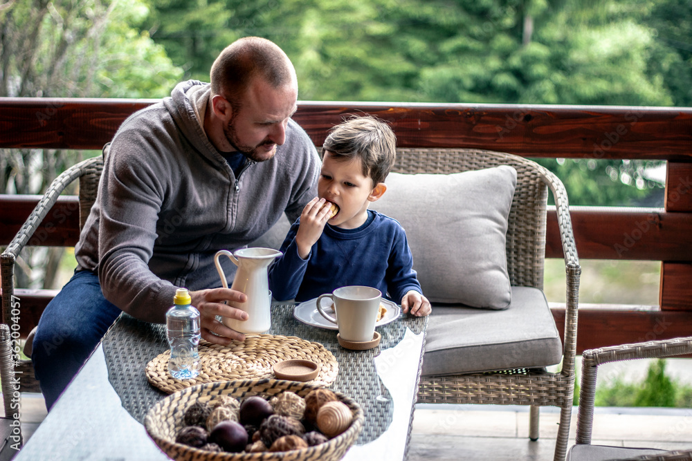 Dad is pouring milk for his cute son in white cup. A beautiful boy looking at his father for making a drink for him. Smiling Son and Father Have Breakfast on the terrace at home. Family bonding. Dad is pouring milk for his cute son in white cup. A beautiful boy looking at his father for making a drink for him. Smiling Son and Father Have Breakfast on the terrace at home. Family bonding.