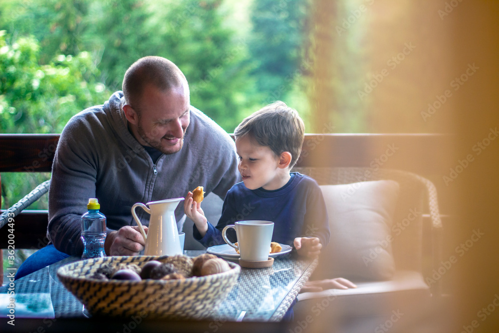 Father and boy talking and smiling while having meal. Todler holding croissant and laughing at his father. Family sitting on terrace and having fun at breakfast. Portrait through the glass. Father and boy talking and smiling while having meal. Todler holding croissant and laughing at his father. Family sitting on terrace and having fun at breakfast. Portrait through the glass.
