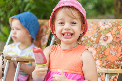 Vacation, summer, village, childhood, sweets, family, leisure, generosity concept. Kids eat popsicles. Brother and sister with fruit ice cream wears hat sitting in the yard outside photo