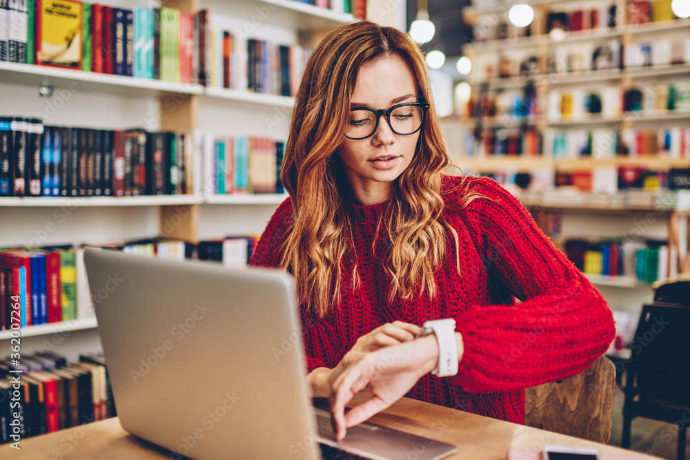 Young skilled serious hipster girl in stylish spectacles for vision correction checking time on modern smartwatch sitting at desktop with laptop computer and waiting end of remote work for today Young skilled serious hipster girl in stylish spectacles for vision correction checking time on modern smartwatch sitting at desktop with laptop computer and waiting end of remote work for today